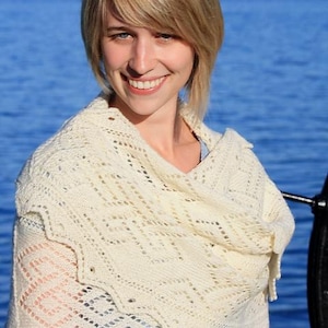 Puede incluir: Chal de punto color crema con un diseño geométrico calado. El chal tiene un borde festoneado y está drapeado sobre los hombros. El fondo muestra una masa de agua y una persona.