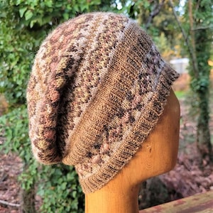 May include: A hand-knitted beanie hat in shades of brown, beige, and pink. The hat features a textured pattern and a folded brim. The hat is displayed on a wooden head form.