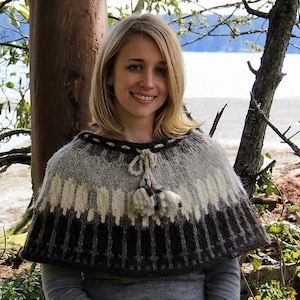 May include: A hand-knitted cape in shades of brown, cream, and gray. The cape features a ribbed bottom edge and a decorative tassel. The cape is worn over a gray long-sleeved shirt. The cape is a cozy and stylish accessory.