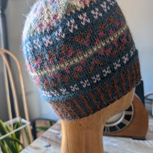 Op de afbeelding: Een handgebreide beanie-muts met een kleurrijk geometrisch patroon. De muts heeft een mix van teal, bruin, roze en crèmekleurig garen. De rand is donkerbruin en de muts wordt gepresenteerd op een houten hoofd.