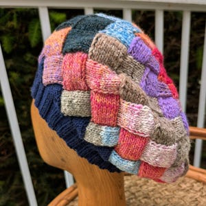 May include: A colorful, hand-knitted beret with a patchwork design. The hat features a dark blue ribbed brim and a top composed of woven squares in shades of pink, purple, orange, blue, and gray. The beret is displayed on a wooden head form.