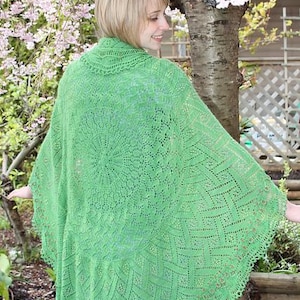 Può includere: Uno scialle verde brillante, a trama aperta, con intricati motivi in pizzo. Lo scialle ha un design circolare al centro e un bordo smerlato. Viene indossato all'aperto, con uno sfondo di alberi in fiore.