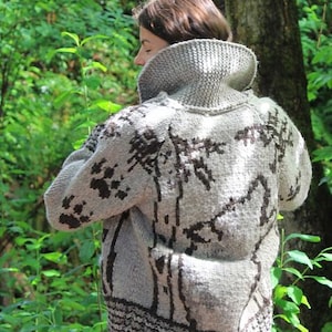 Puede incluir: Un suéter de punto gris con cuello alto y un diseño de árbol marrón oscuro. El suéter tiene un aspecto texturizado y un dobladillo acanalado. La persona que lleva el suéter está en un entorno forestal.