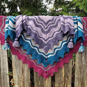 May include: A handmade crochet shawl in shades of purple, blue, and white, with a scalloped edge and tassel accents. The shawl is draped over a wooden fence, showcasing its intricate design and color palette.