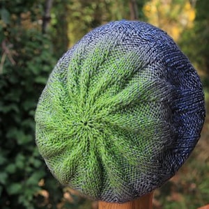 Op de afbeelding: Een handgebreide beanie muts met een spiraalvormig ontwerp. De muts heeft een kleurovergang van levendig groen in het midden naar tinten blauw en grijs aan de bovenkant. De muts wordt getoond op een houten standaard, die de textuur en kleurverloop benadrukt.