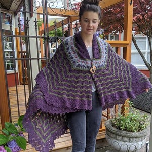 May include: A handmade purple and green knitted shawl with a unique design. The shawl features a decorative wooden clasp and a patterned collar. The shawl is worn by a person in the image.