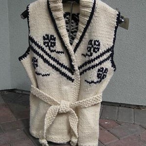 May include: Cream-colored knitted vest with a shawl collar and a tie belt. The vest features a black clover-like pattern and black trim. The garment is displayed on a hanger against a neutral background, showcasing its design and texture.