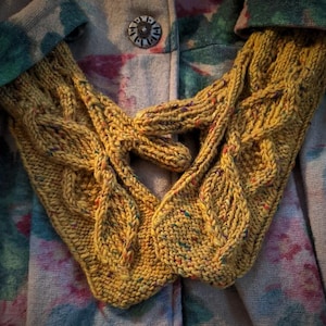 Puede incluir: Un par de mitones de punto amarillo mostaza con un diseño de trenzas y detalles en forma de hojas. Los mitones, salpicados con motas de colores, se muestran sobre un abrigo con estampado floral. Un accesorio acogedor.