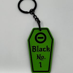 May include: A green coffin-shaped keyring with the text "Black No. 1" in black gothic font. The keyring has a black ring and chain. The top of the coffin shape has a black horizontal line.