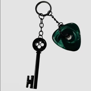 Pode incluir: Um chaveiro preto com uma chave e um pingente verde em forma de coração. A chave é preta com um topo circular e uma haste longa e retangular. O pingente é verde escuro com um padrão de redemoinho. O chaveiro tem um anel preto.