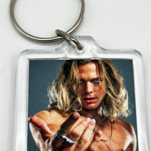 Könnte beinhalten: Transparenter Acryl-Schlüsselanhänger mit einem quadratischen Bild einer Person mit langen, welligen blonden Haaren und blauen Augen. Die Person ist ohne Hemd und hat einen silbernen Ring am Finger. Oben ist ein silberner Schlüsselring befestigt.