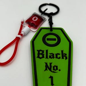 Könnte beinhalten: Ein grüner Sarg-Schlüsselanhänger mit dem Text "Black No. 1" in Schwarz. Der Schlüsselanhänger hat einen schwarzen Rand und einen schwarzen Ring. Ein roter und weißer Blutbeutel-Anhänger ist befestigt.