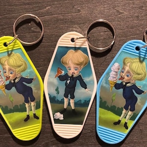 May include: Three keychains with a retro design. Each keychain features a cartoon illustration of a young person in a dark blue outfit. One keychain is yellow, one is white, and one is blue. The young person is holding an ice cream cone or a small bird.
