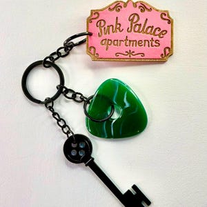 Può includere: Un portachiavi con un cartello rosa con la scritta "Pink Palace apartments" in oro. Ha un ciondolo triangolare verde marmorizzato, una chiave nera e maglie di catena nere.