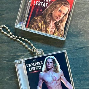 Op de afbeelding: Twee doorzichtige plastic sleutelhangers met afbeeldingen uit "The Vampire Lestat". Elke sleutelhanger heeft een zilveren kogelketting. De afbeeldingen tonen een man met lang blond haar en de filmtitel.