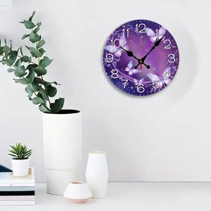May include: A round wall clock with a purple and white butterfly design. The clock face features white numbers and black clock hands. The clock is mounted on a white wall.