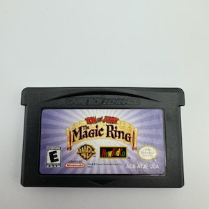 Op de afbeelding: Zwarte Game Boy Advance-cartridge met "Tom and Jerry: The Magic Ring." Het label bevat het Warner Bros.-logo, NewKidCo en de ESRB-classificatie "E" voor iedereen. De cartridge is gelicentieerd door Nintendo.