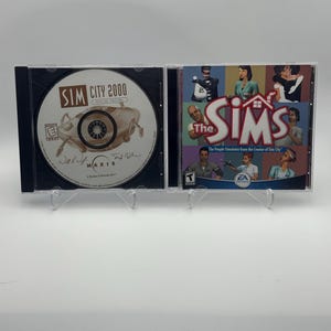 Puede incluir: Dos cajas de CD de plástico transparente una al lado de la otra. La caja de la izquierda contiene un disco de SimCity 2000 Special Edition con un gráfico blanco y negro. La caja de la derecha muestra la portada del juego The Sims con personajes.