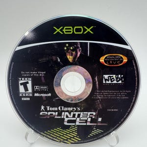 May include: An Xbox game disc for Tom Clancy's Splinter Cell, featuring a character with night-vision goggles. The black disc includes the ESRB Teen rating, Dolby Digital logo, Microsoft branding, and a Download New Content on Live emblem.