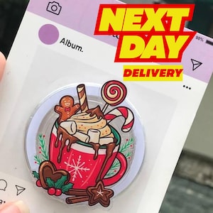 May include: A clear phone grip with a festive design featuring a red mug of hot chocolate with marshmallows, a gingerbread man, and a candy cane. The words "NEXT DAY DELIVERY" are displayed in yellow. The background has social media icons.