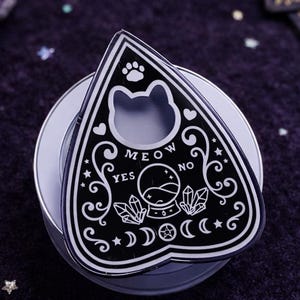 May include: A black and white planchette with a cat-shaped cutout and the words "MEOW YES NO." The planchette is decorated with moons, stars, and crystals. It sits on a white circular base, with a dark purple background.