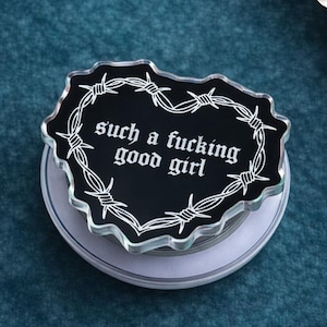 May include: A black heart-shaped phone grip with white barbed wire detailing and the phrase "such a fucking good girl" in gothic font. The grip is on a white and silver base, set against a teal background.