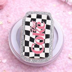 May include: A Diet Coke-themed wireless phone charger. The charger has a rectangular shape with a black and white checkered pattern and the words "Diet Coke" in pink. The charger is surrounded by a silver glitter border.