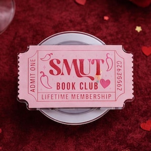 May include: A pink ticket-shaped item with the words "SMUT BOOK CLUB LIFETIME MEMBERSHIP" printed on it. The ticket is decorated with chili pepper and heart graphics. The background is a deep red with scattered heart and star-shaped confetti.