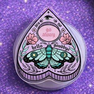 May include: A clear, heart-shaped planchette with a decorative design featuring a moth, flowers, and the words "hello" and "goodbye." The planchette also has the words "yes," "no," and "go away." The design is in shades of purple, teal, and pink.