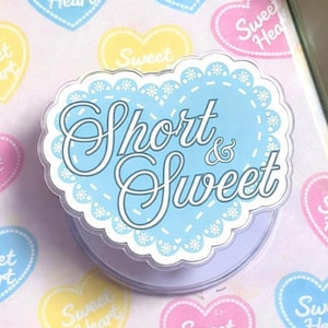 May include: A light blue phone grip with the words "Short & Sweet" in white script. The grip has a scalloped edge and is surrounded by heart-shaped stickers that say "Sweet Heart" in various colors: pink, yellow, and blue.