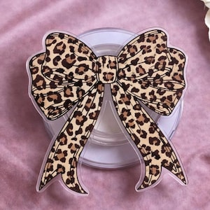 May include: A clear, leopard print bow-shaped accessory. The bow features a tan and brown spotted pattern. The accessory is placed on a clear, circular base, set against a pink background. The bow is a decorative item.