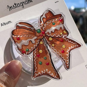 Puede incluir: Un soporte transparente para teléfono con un diseño de lazo de pan de jengibre. El lazo es marrón con detalles de glaseado blanco y chispas de colores. El logotipo de Instagram y la palabra "Album" son visibles en el fondo.