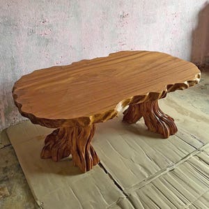May include: A handcrafted wooden coffee table with a natural edge design. The table features a light brown, oval-shaped top supported by three intricately carved tree trunk-style legs. The table measures approximately 122 cm long and 61 cm wide.