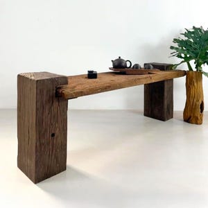 May include: A weathered wooden bench with a natural, rustic aesthetic. The bench has a thick, rectangular top supported by two dark brown wooden legs. A small tea set sits on the bench, alongside a wooden tray and a decorative vase with green leaves.