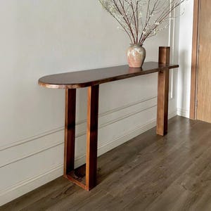 Puede incluir: Una consola larga de madera marrón oscuro con un borde frontal redondeado. La mesa tiene dos patas en forma de U en un lado y una sola pata rectangular en el otro. Un jarrón con flores blancas y un pequeño bloque de madera encima.