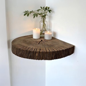 Op de afbeelding: Een rustiek, halfrond houten plankje aan een witte muur. De plank is gemaakt van een dwarsdoorsnede van een boomstam, met de natuurlijke nerf en textuur. Twee brandende kaarsen en een glazen vaas met groen staan op de plank.