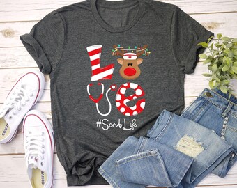 May include: Dark grey t-shirt featuring a medical-themed "LOVE" design with a reindeer wearing a nurse's cap and the text "#ScrubLife". The image also shows white trainers and blue denim jeans.