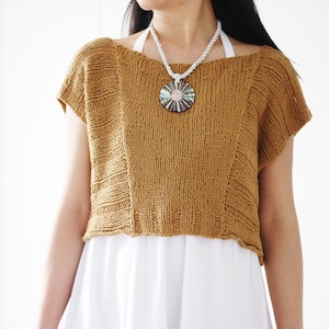 Puede incluir: Un top corto de punto de color mostaza con escote amplio y mangas cortas. El top tiene un corte ligeramente holgado y un patrón de punto texturizado. La modelo lleva un collar blanco con un gran colgante redondo.