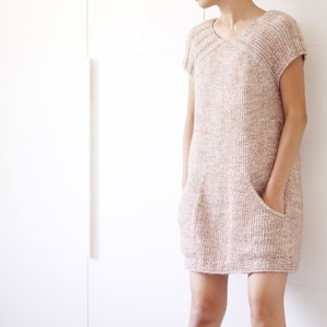 Könnte beinhalten: Ein locker sitzendes, kurzärmeliges Strickkleid in einem hellrosa und weißen, gesprenkelten Muster. Das Kleid hat einen runden Ausschnitt und zwei aufgesetzte Taschen.