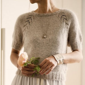 May include: A light grey knitted top with short sleeves and decorative shoulder patterns. The model is wearing a white skirt and holding a pink flower. A silver necklace and bracelet complete the look. The overall aesthetic is soft and feminine.