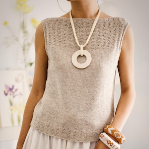 May include: A beige knitted sleeveless top with a matching necklace featuring a large circular pendant. The outfit includes a white skirt and two patterned bracelets. The overall aesthetic is minimalist and natural.