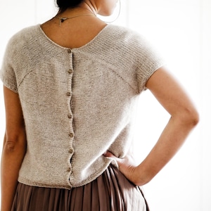 Può includere: Un top di maglia beige con maniche corte e chiusura con bottoni sul retro. Il top è indossato sopra una gonna plissettata marrone.