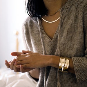 Può includere: Una donna indossa un maglione di lana marrone chiaro con scollo a V e nappe. Indossa una collana a catena dorata e un braccialetto dorato.