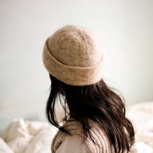 Può includere: Una persona che indossa un berretto di lana marrone e un gilet di lana color crema. La persona è seduta su un letto bianco.