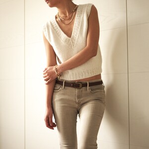 May include: A white sleeveless knitted sweater with a v-neckline and a brown leather belt. The sweater is worn over a pair of light brown jeans.