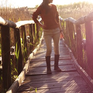 May include: A person wearing a brown sweater and jeans walks on a wooden bridge over a grassy area. The bridge is made of wooden planks and has railings on both sides. The person is wearing brown boots and has long dark hair.