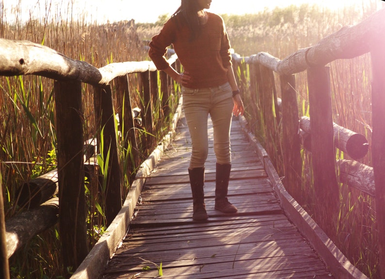 May include: A person wearing a brown sweater and jeans walks on a wooden bridge over a grassy area. The bridge is made of wooden planks and has railings on both sides. The person is wearing brown boots and has long dark hair.