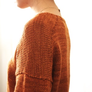 May include: A close-up of a person wearing a rust-colored knitted sweater with a textured stitch pattern. The sweater has a round neckline and long sleeves.