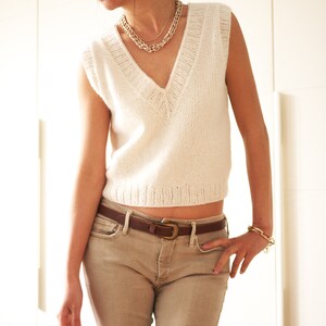 May include: A white sleeveless knitted sweater with a v-neckline and a brown leather belt. The sweater is cropped and the belt is fastened at the waist.