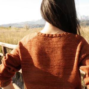May include: A woman wearing a brown knitted sweater stands on a wooden walkway overlooking a field of tall grass. The background features a mountain range in the distance.